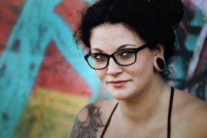 A woman with dark hair and glasses looking at the camera. She has a mandala tattoo on her shoulder and is stood against a multicoloured backdrop that looks like street art paintings.