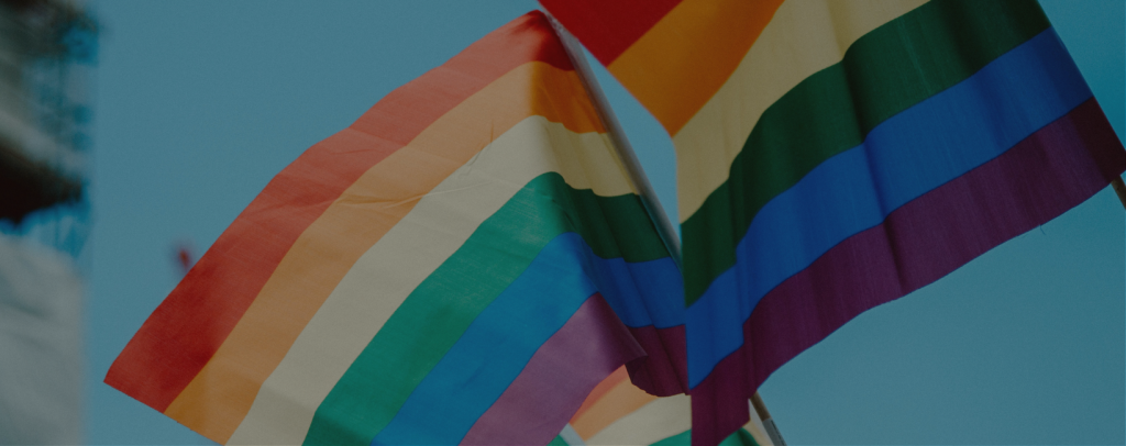 Two rainbow pride flags fluttering in the breeze against an unfocused backdrop of a blue sky.